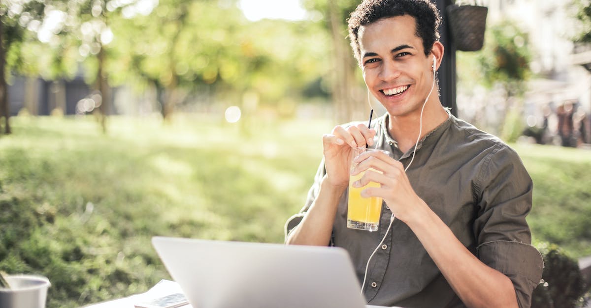 cheerful-guy-with-laptop-and-earphones-sitting-in-park-while-drinking-juice-and-smiling-at-camera-9134710