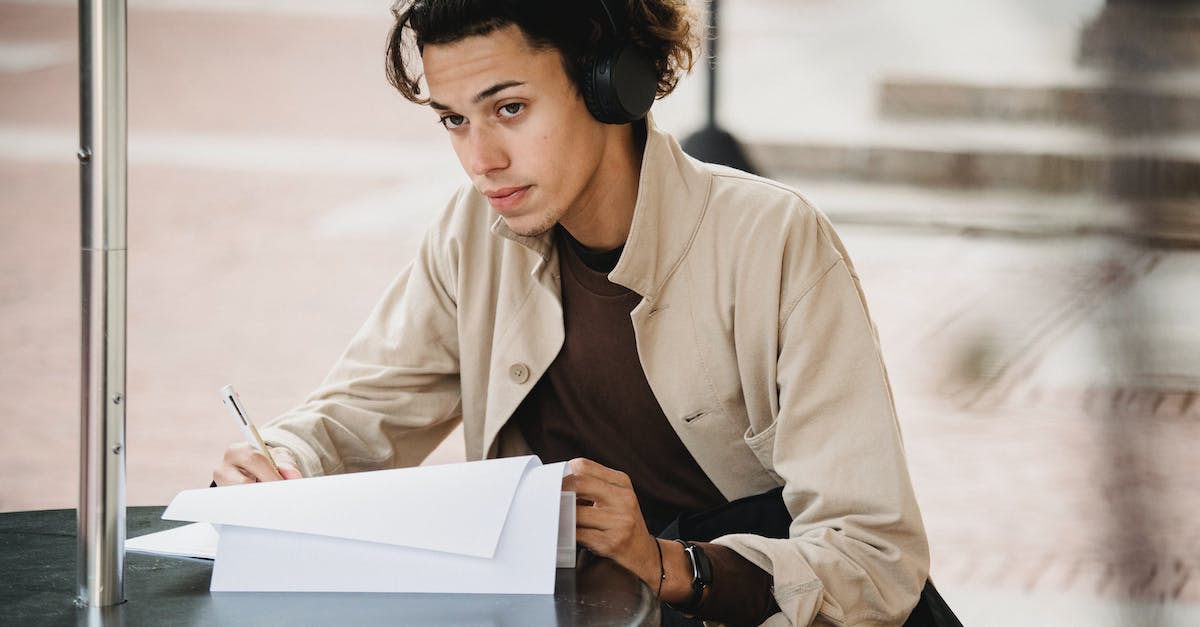 concentrated-male-student-in-wireless-headphones-sitting-at-table-and-writing-answers-in-document-du-7059604