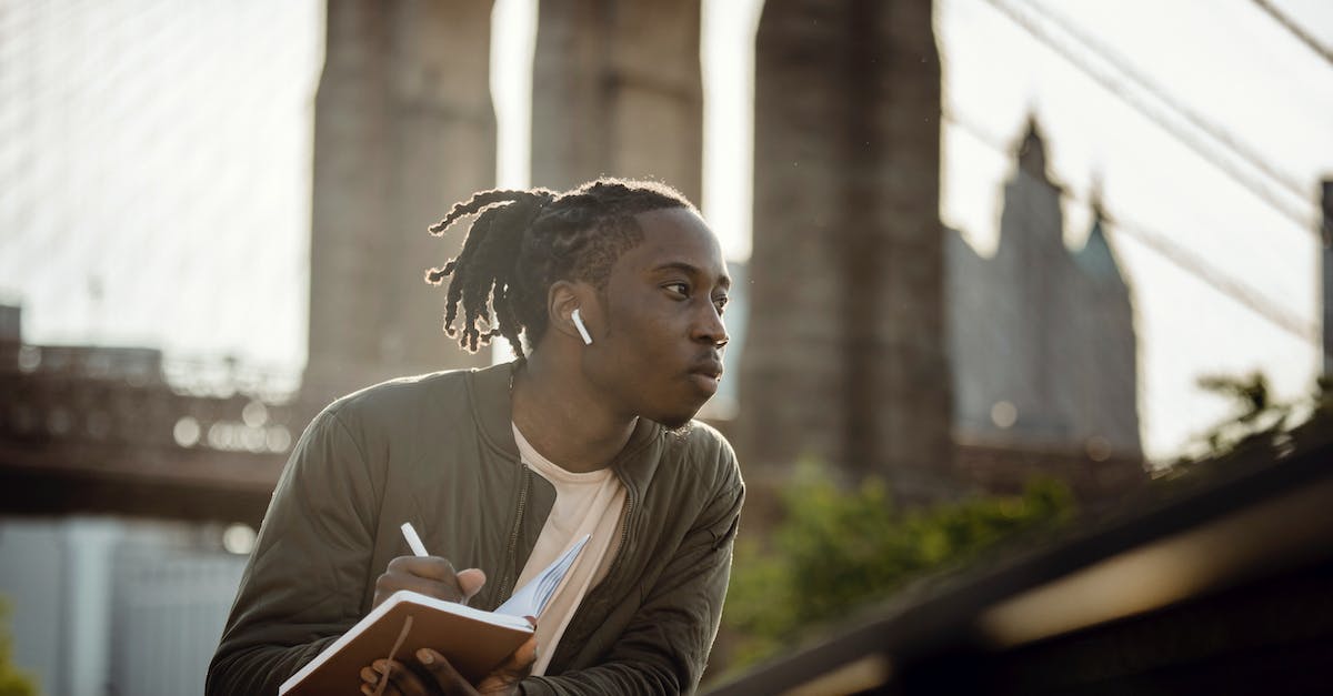 positive-black-man-listening-to-music-with-wireless-earphones-and-taking-notes-in-diary-on-street-7103548