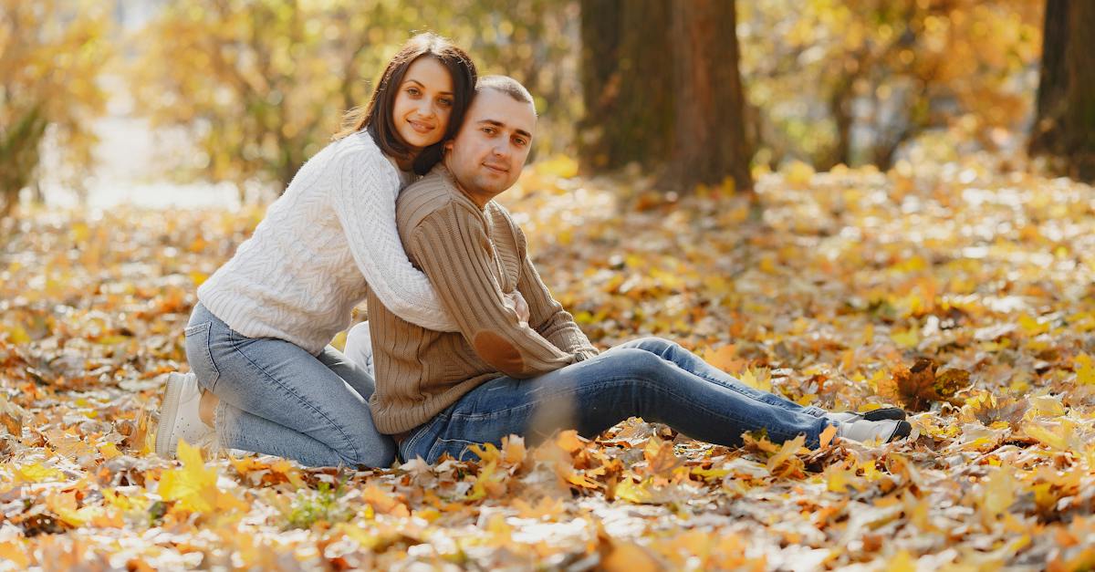 side-view-of-cheerful-girlfriend-in-white-sweater-sitting-on-lawn-with-fallen-yellow-leaves-hugging-3697856